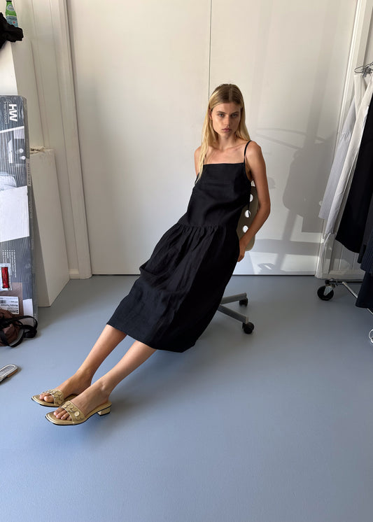 A woman with long blonde hair wears the Gathered Halter Dress - Black, styled with beige heeled sandals as she sits on a chair in a minimal, light-filled room featuring stone-washed French linen, a pale blue floor, and white walls.