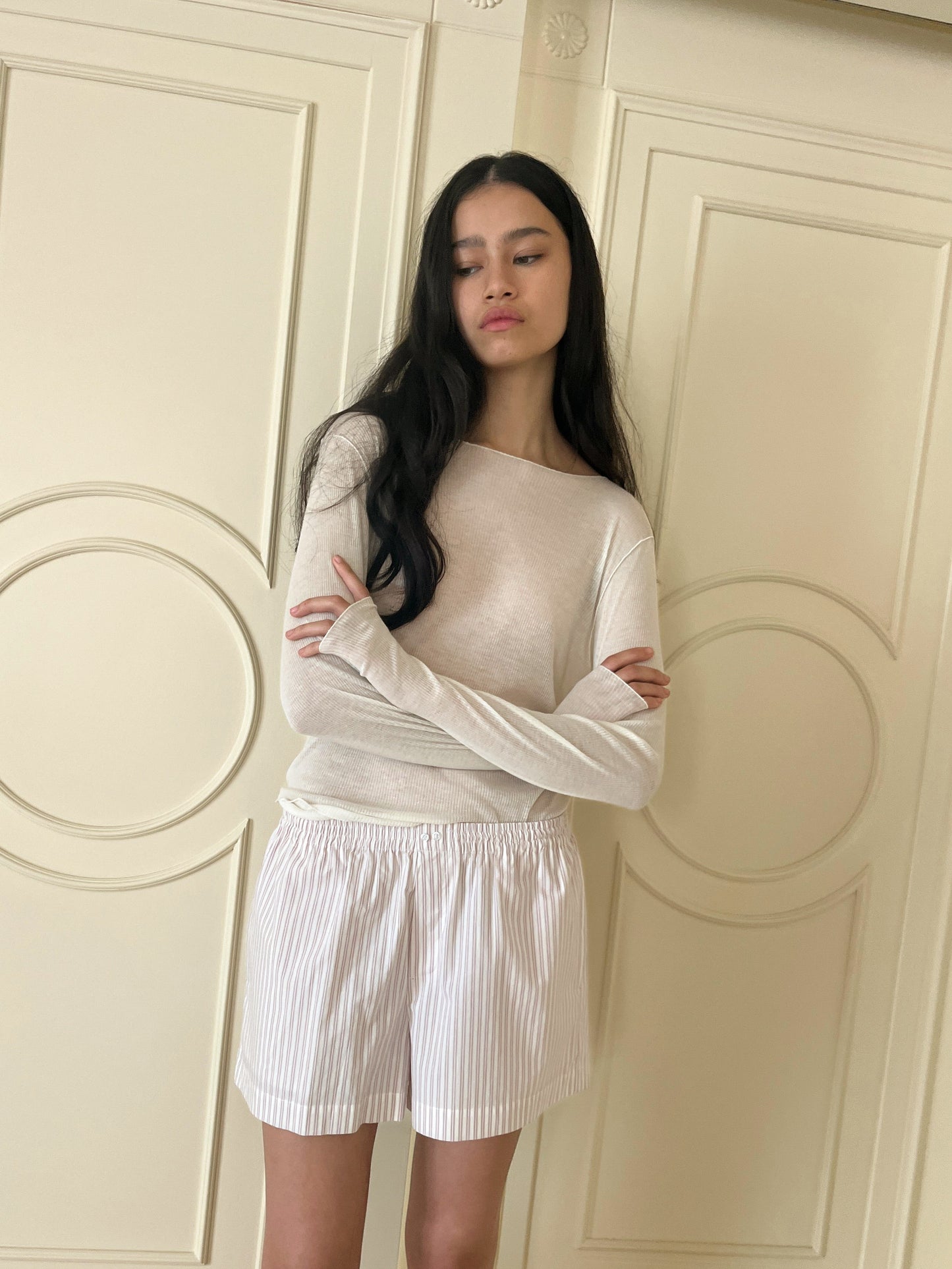 A woman with long dark hair stands before cream-paneled doors, arms crossed and calm, wearing a light beige top and the Organic Cotton Boxer - Musk Stripe, featuring white fabric with stripes and an elastic waistband.