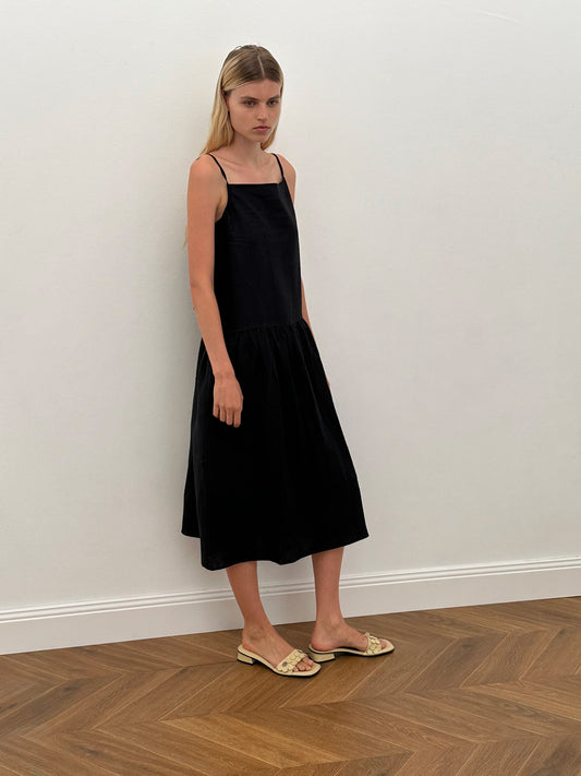 A woman with straight blonde hair wears the Gathered Halter Dress - Black, a sleeveless, loose-fitting halter style in stone-washed French linen, paired with beige flat sandals against a plain white wall on a herringbone wood floor.