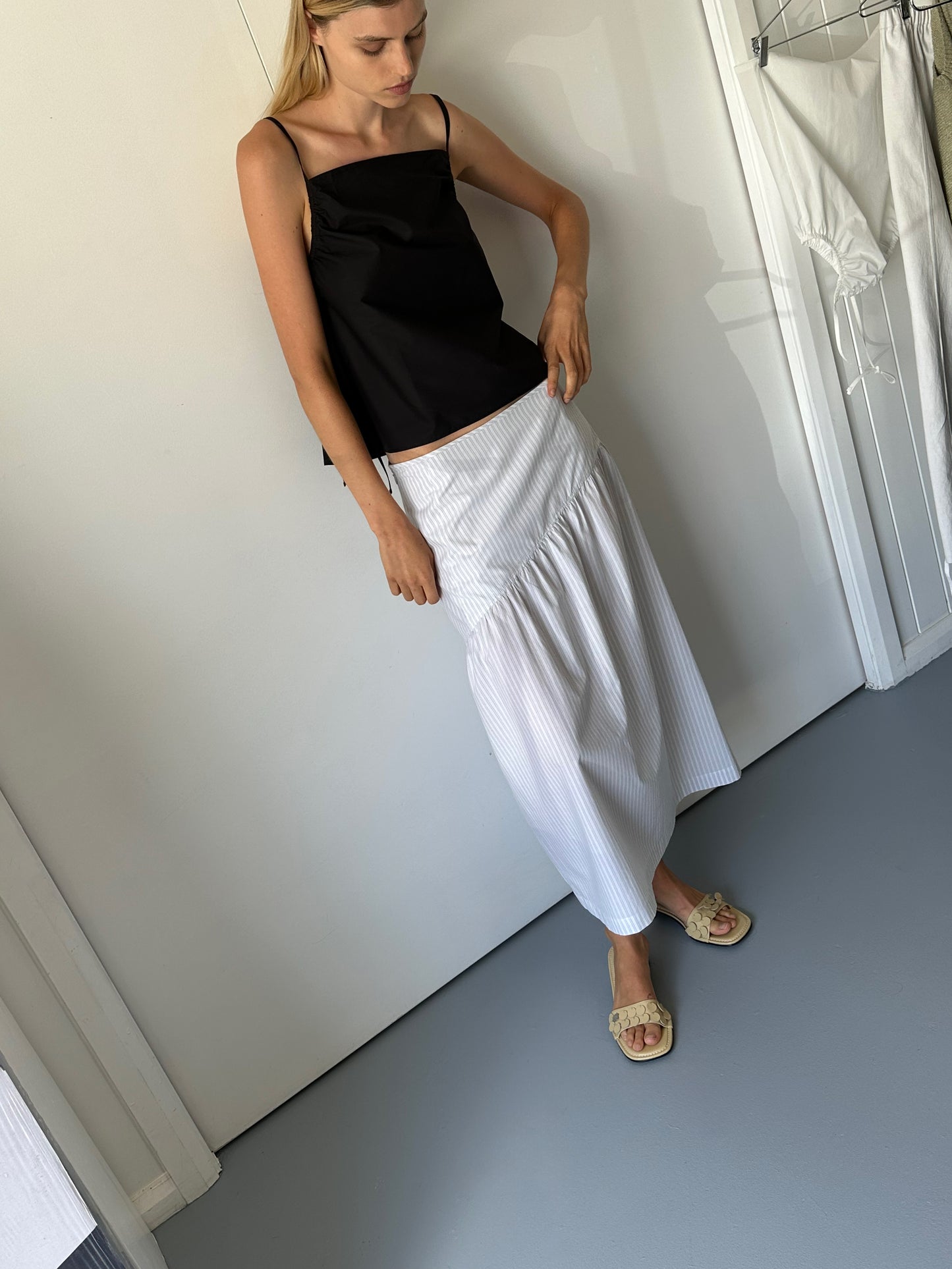 A woman stands indoors wearing a black sleeveless top, beige sandals, and the 0 Angle Skirt - Sage Stripe. She leans slightly forward, looking down with one hand resting on her skirt.