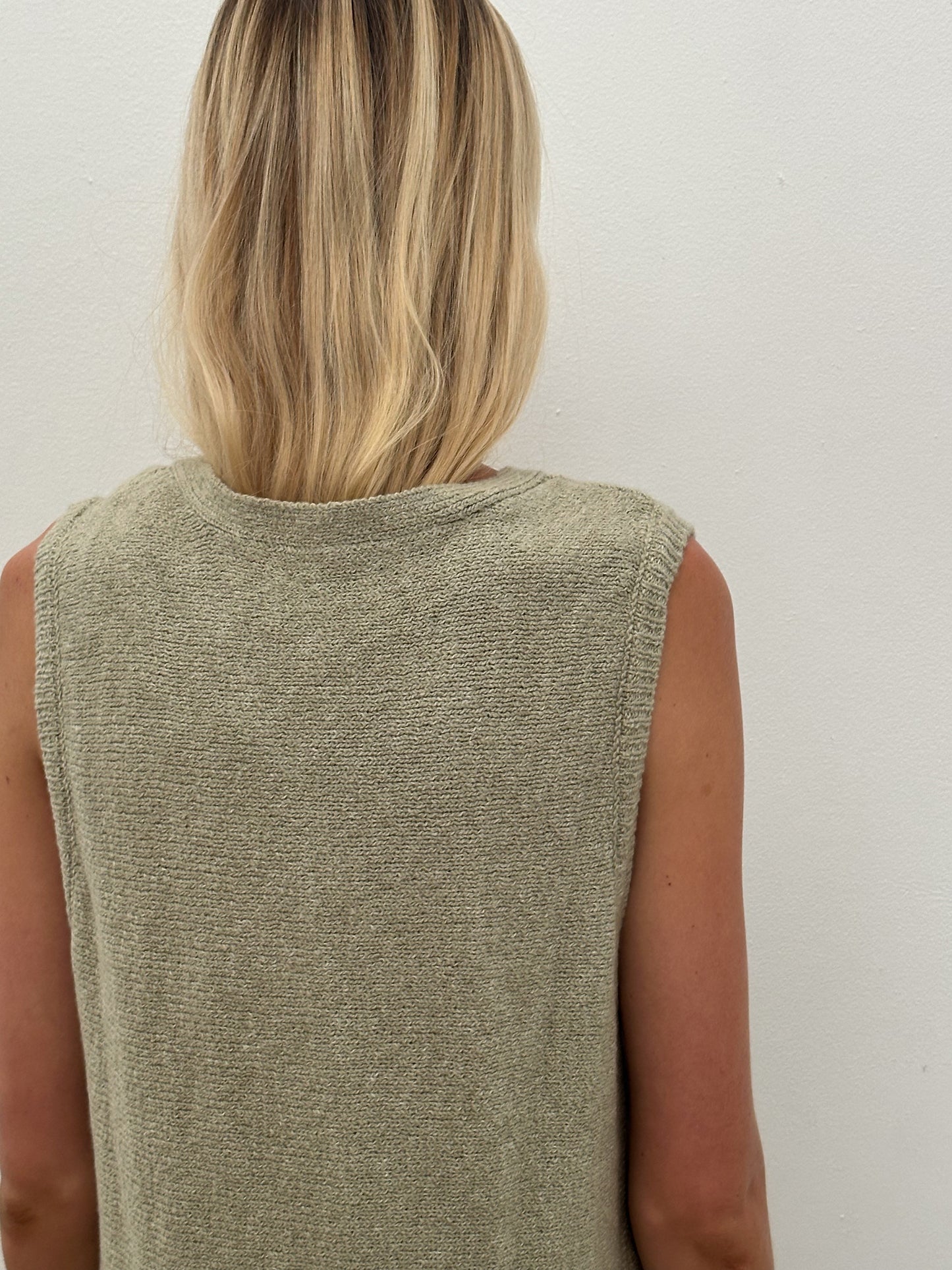 A person with shoulder-length blonde hair is seen from behind wearing the 0 Knit Vest - Powered Sage, a lightweight knit in light olive green, against an off-white background.