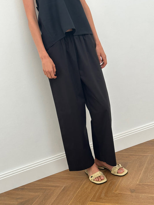 A person stands indoors wearing the Ease Trouser - Black from Deiji Studios, paired with a black sleeveless top and light-colored ruffle-detail sandals. Only the lower half of their body is visible, with arms relaxed by their sides.