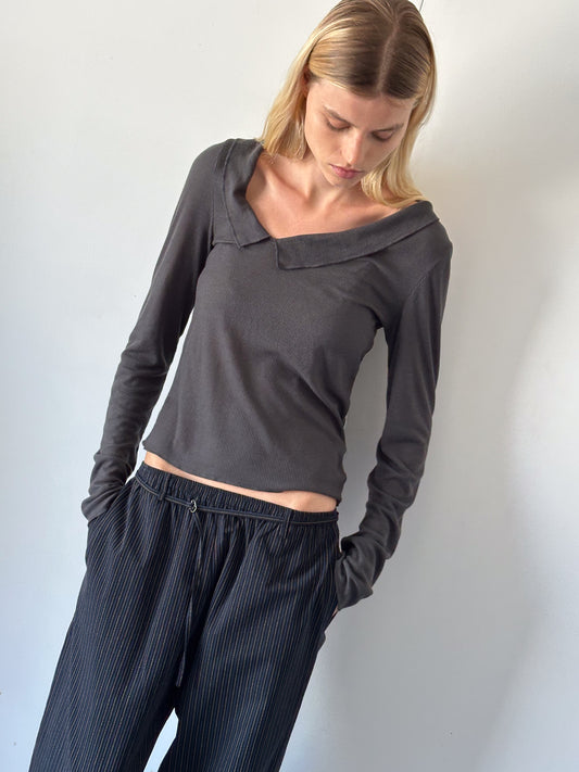 A person with straight blonde hair looks down, wearing the Cross Collar Top - Night Blue with an asymmetrical neckline and loose, pinstriped black pants, standing against a plain light-colored wall.