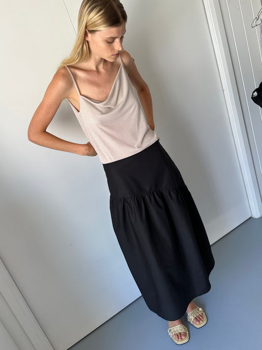 female model wears the split top by Deiji Studios in shell
a sheer fine knit relaxed tank with a fold over cowl neckline and low draping back, features self fabric rouleau straps, baby lock hem finishes and a asymmetric bottom detail
made from a responsible blend of 85% Recycled Polyester and 15% Viscose
paired with a black cotton skirt