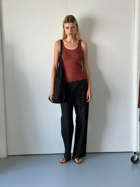 A woman stands indoors against a white wall, wearing the Knit Tank - Crimson with a racerback, loose black pants, sandals, and carrying a black shoulder bag. Her blonde hair is down in a minimal, modern setting.