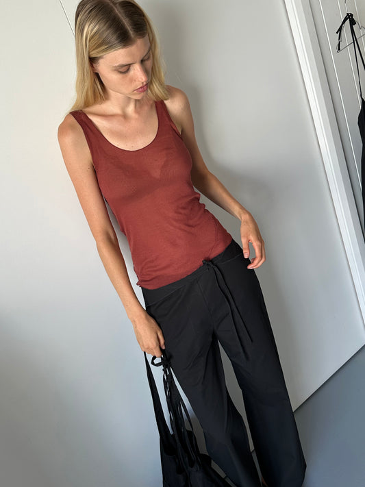 A woman with straight blonde hair wears the Knit Tank - Crimson with a racer-style back and loose black pants, standing indoors with white walls and a door, looking down while holding a black bag.