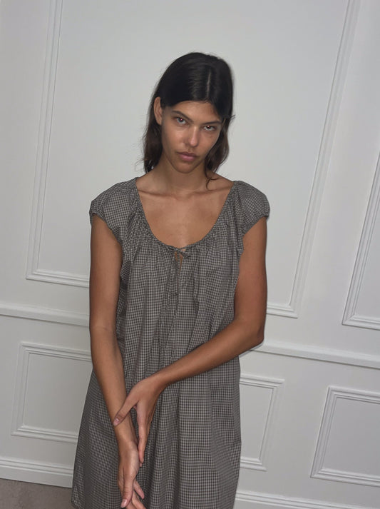 A woman with long dark hair wears the Capped Sleeve Dress - Khaki Check, standing before a white paneled wall and gazing at the camera with a neutral expression, her hands folded gently in front of her.