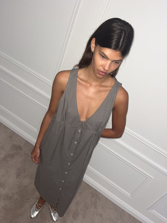 A woman with long dark hair stands indoors against a white paneled wall, wearing the Tuck Tie Dress—a sleeveless, deep V-neck, button-front dress with a checkered pattern—and silver pointed-toe shoes. She looks slightly downward.