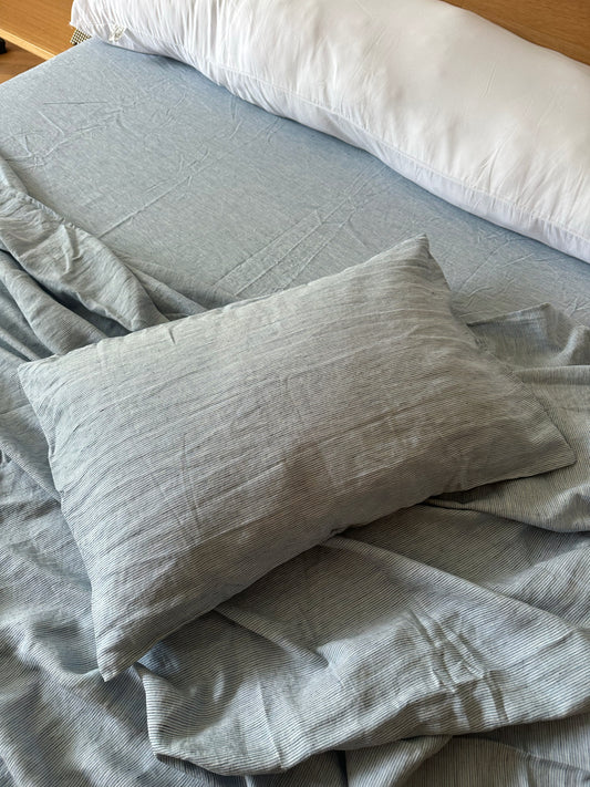 A bed with light gray striped pillow slips and a matching pillow, topped with a white bolster at the headboard. The bedding looks slightly rumpled.