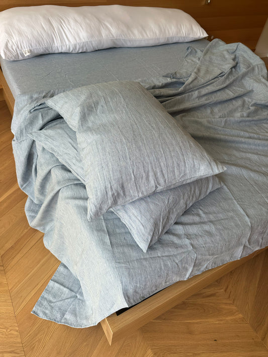 A bed featuring the Sheets - Blue Stripe set, slightly wrinkled and partially unmade, rests on a wooden floor with a light brown headboard and white pillow—styled by Deiji Studios.
