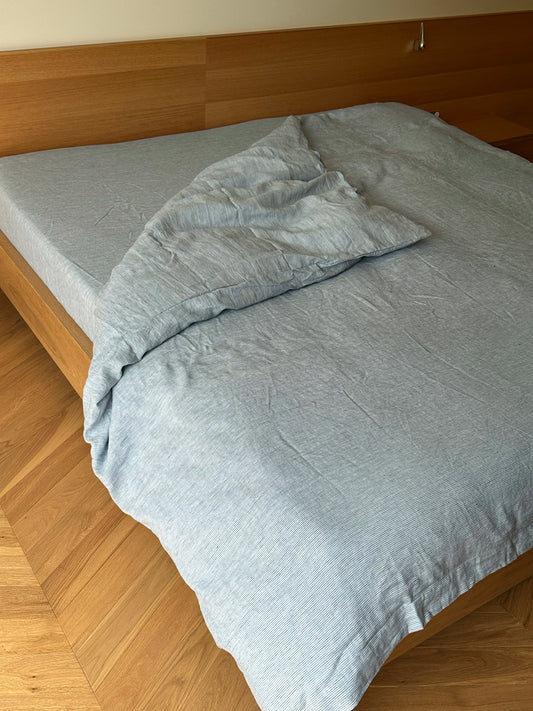 A wooden bed with linen sheets is topped with the Blue Stripe duvet cover and matching pillow, slightly wrinkled and partially unmade, resting on a light wood herringbone floor.