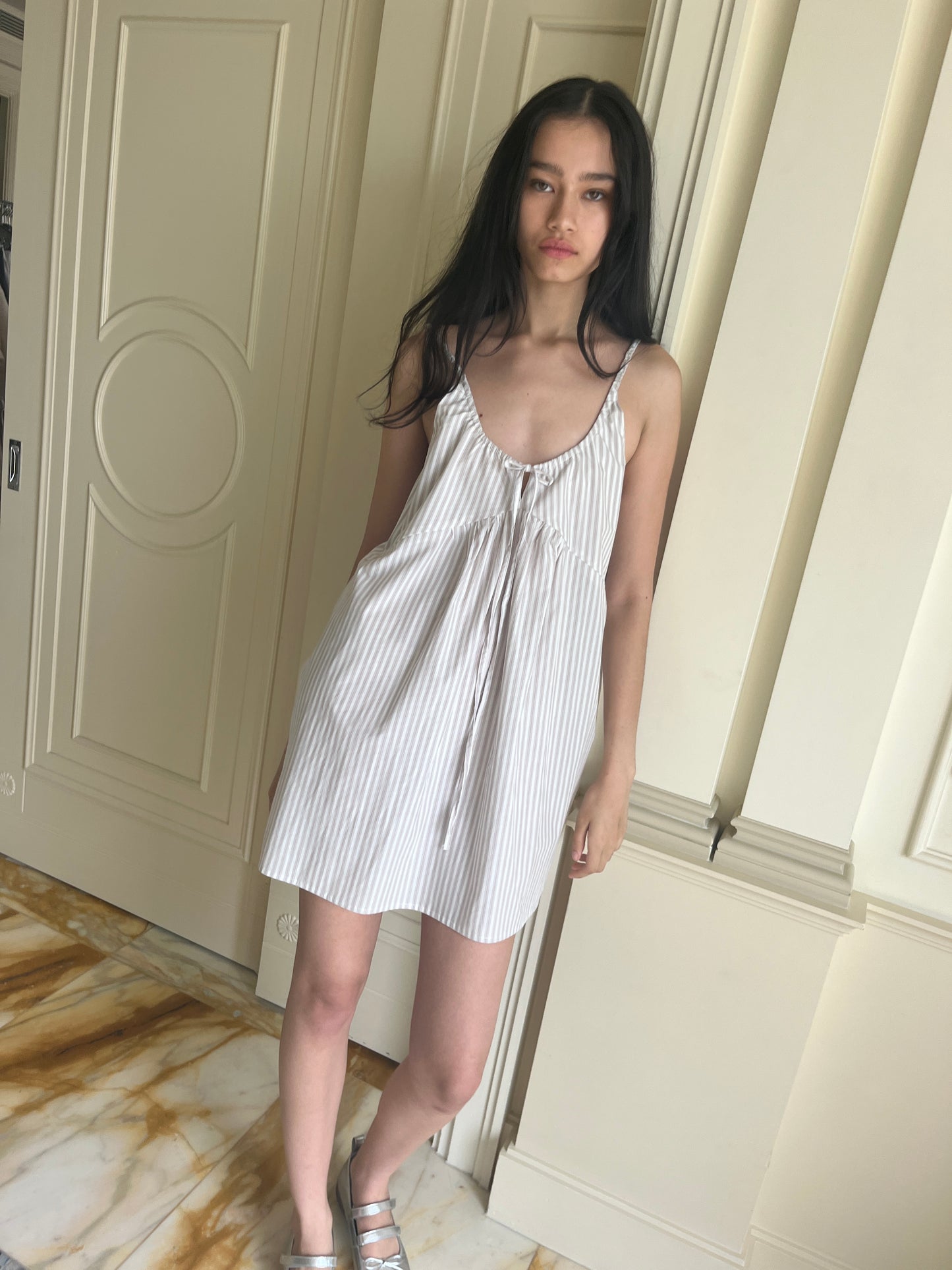 A woman with long dark hair stands indoors wearing the Deiji Studios Double V Dress - Lake Stripe and sandals, posing before cream-paneled doors on a marble floor.