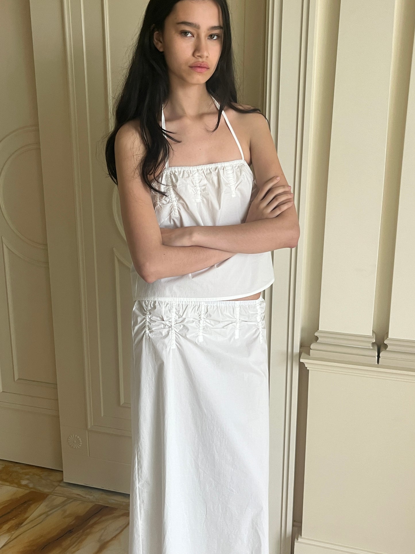 A young woman with long dark hair stands indoors with arms crossed, wearing the Tube Top - White from Deiji Studios, paired with a matching long white skirt, set against a cream-colored, ornately paneled wall.
