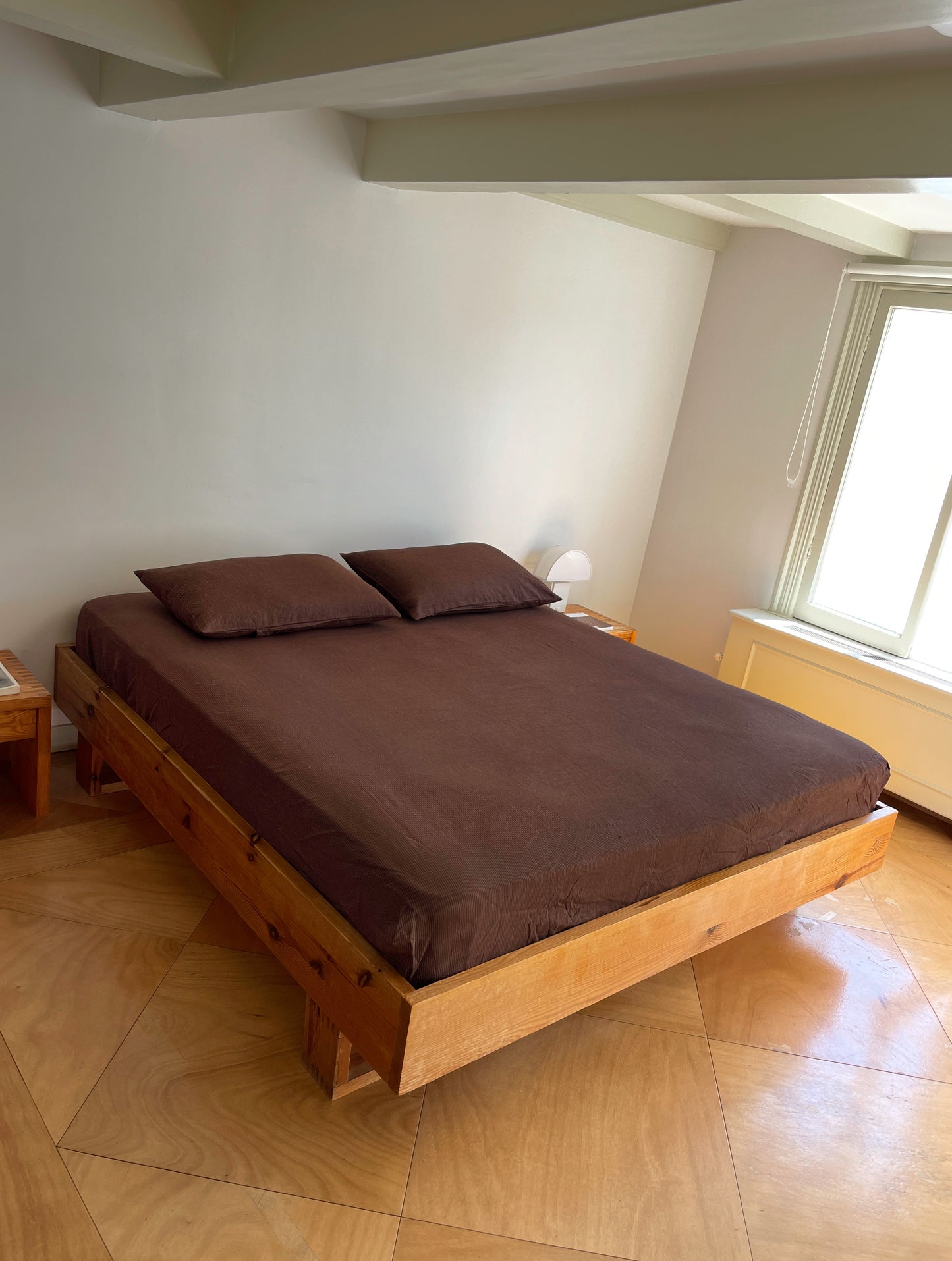 A minimalist bedroom with a wooden bed frame, Sheets - Mahogany Royal Stripe bedding, two matching pillows, light wood floors, a small white lamp on a nightstand, and sunlight streaming through a large window.