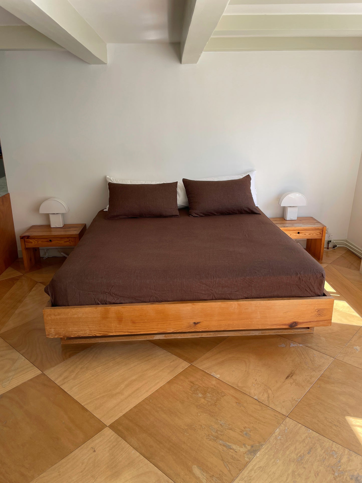 A minimalist bedroom featuring a wooden bed frame, brown bedding and pillows, matching nightstands, small white lamps, and organic French linen Sheets - Mahogany Royal Stripe against a white wall and light wood floor.