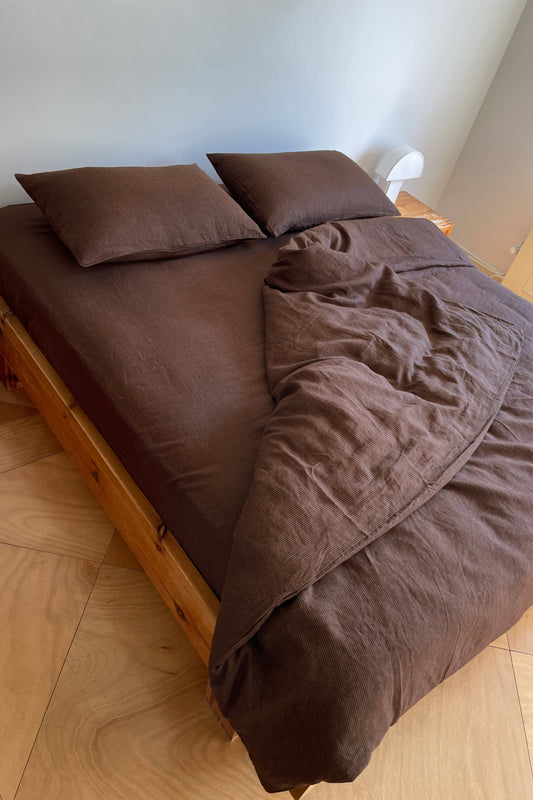 A wooden bed with two brown pillows and a brown duvet, partially unmade, sits in a light-walled room with a wooden floor. A small white lamp and wooden nightstand are beside the bed.