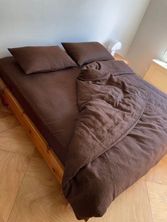 A wooden bed is dressed in Sheets - Mahogany Royal Stripe, with two matching pillows and a brown comforter partially pulled back, all in a sunlit room with pale walls and a small white lamp on the bedside table.