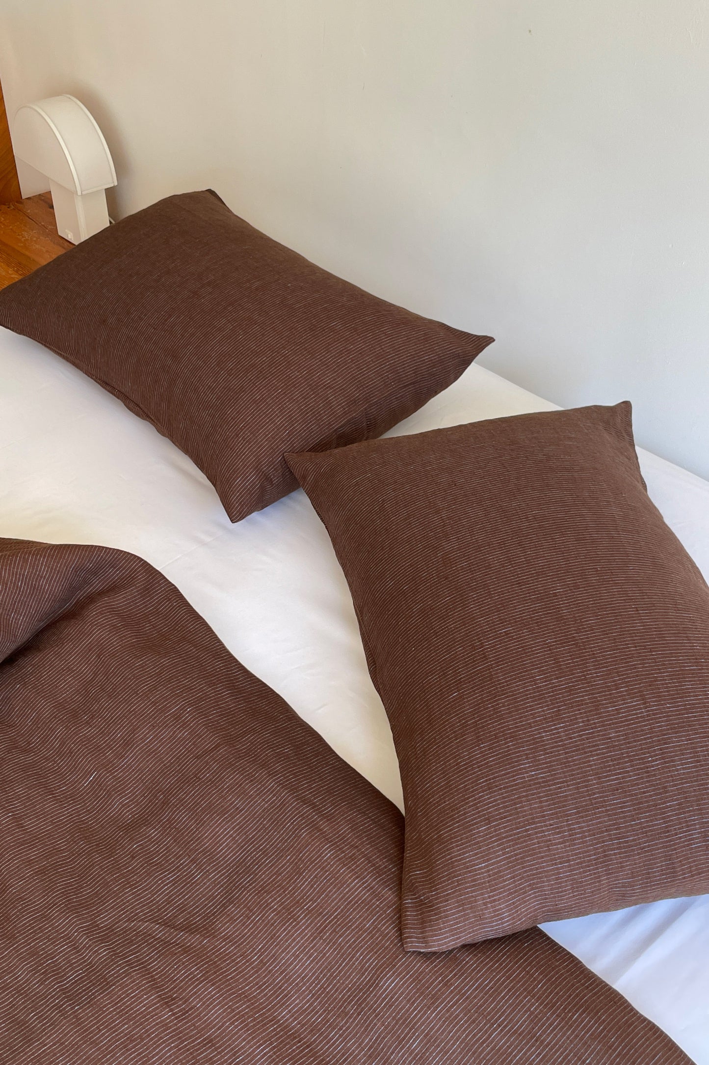 A neatly made bed features two brown Pillow Slips and a matching brown duvet cover on white sheets, beside a wooden nightstand with a small white lamp.