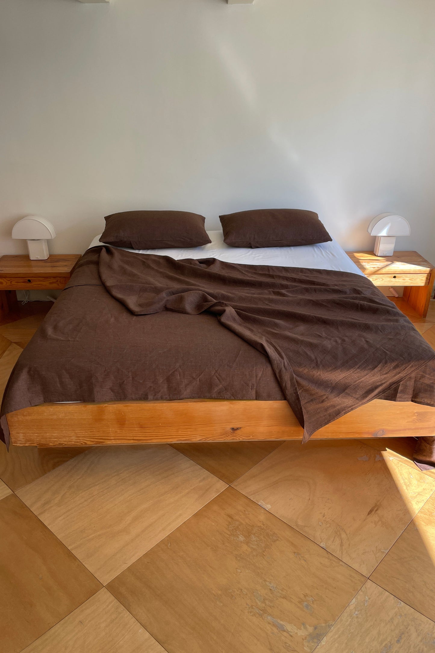 A minimalist, sunlit room with white walls features a wooden bed dressed in Sheets, two pillows, and brown bedding, flanked by small wooden nightstands with white lamps on a light wood floor.