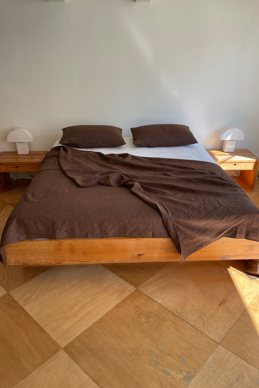 A wooden bed with Sheets - Mahogany Royal Stripe and two pillows stands against a plain white wall. Two small nightstands with white lamps are on either side, and large square tan tiles cover the floor.