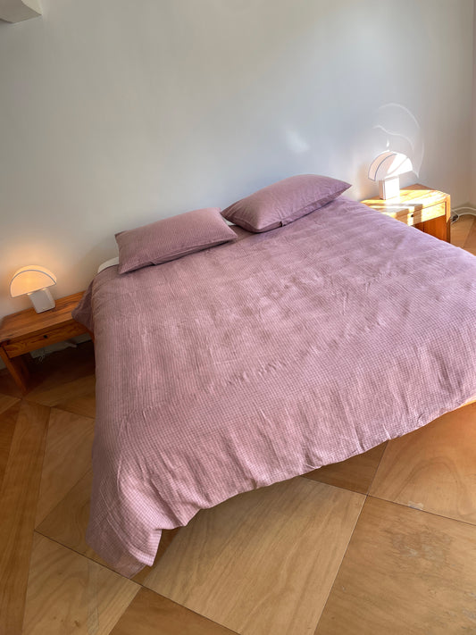 A neatly made bed with the hypo-allergenic, light purple Musk Check duvet cover and two matching pillows sits between wooden nightstands with modern dome lamps. The floor is light wood, and the wall is plain white.
