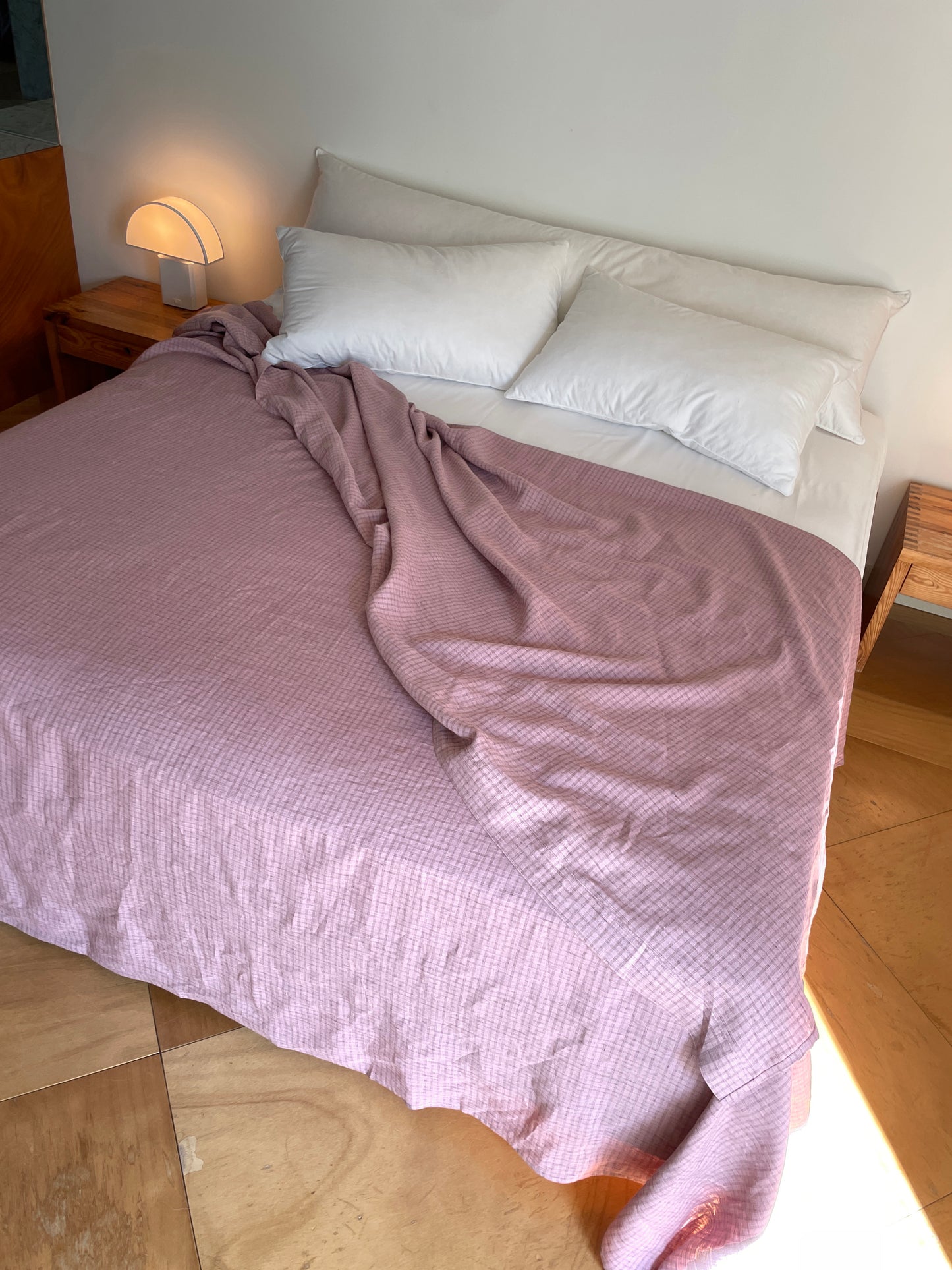 A neatly made bed with three white pillows and a light purple Musk Check sheet, partially folded back, featuring hypo-allergenic bedding, beside wooden nightstands and a small lamp in a sunlit minimalist room.