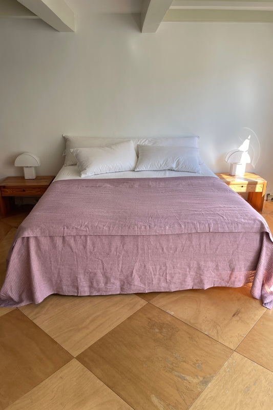 A neatly made bed features white pillows and the Sheets light pink bedspread, centered against a white wall with wooden nightstands and white lamps. The room has light wooden floors and a minimalistic style.