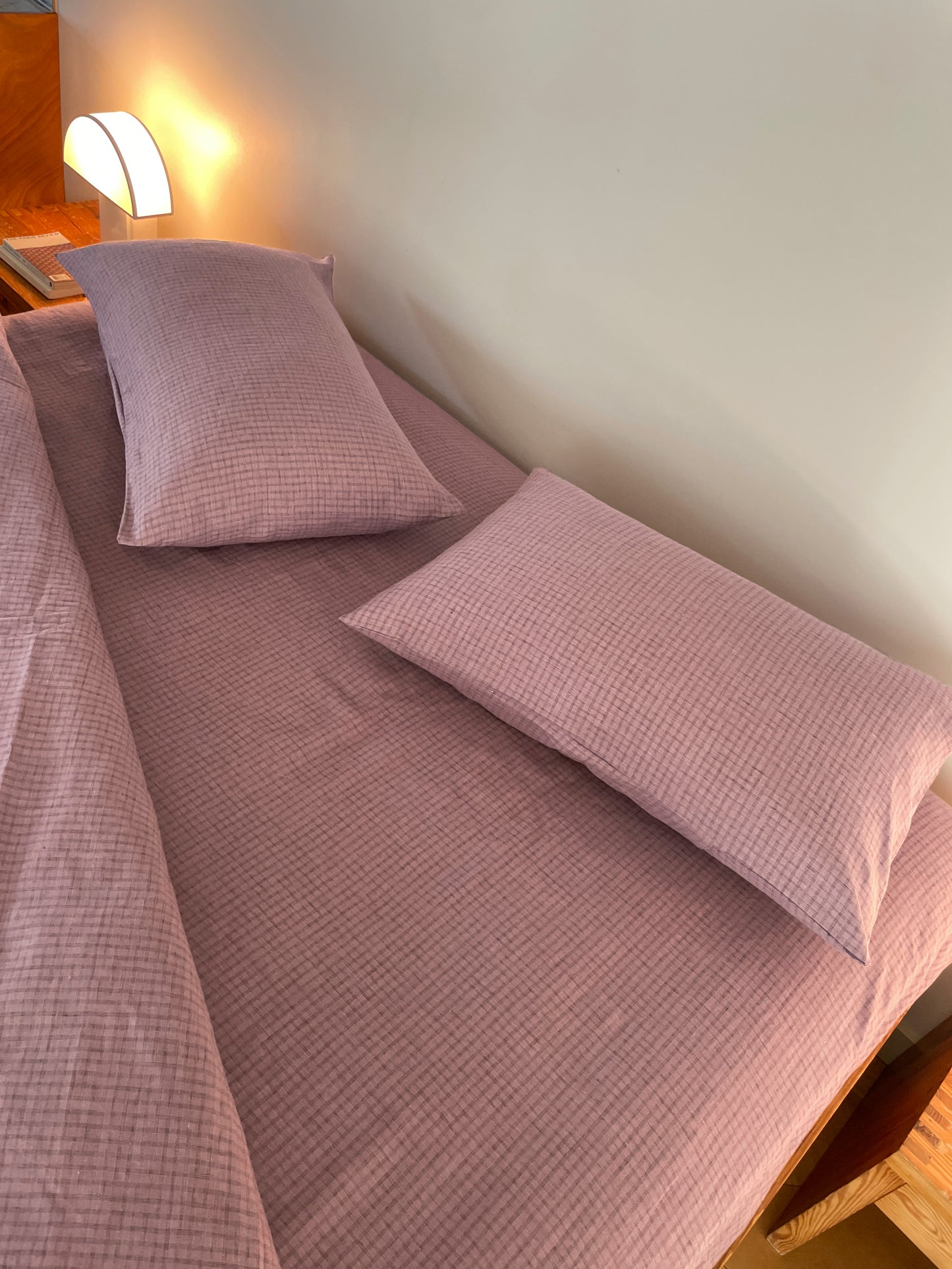 A neatly made bed with Musk Check organic linen sheets and two matching pillows, softly lit by a modern bedside lamp on a wooden nightstand.