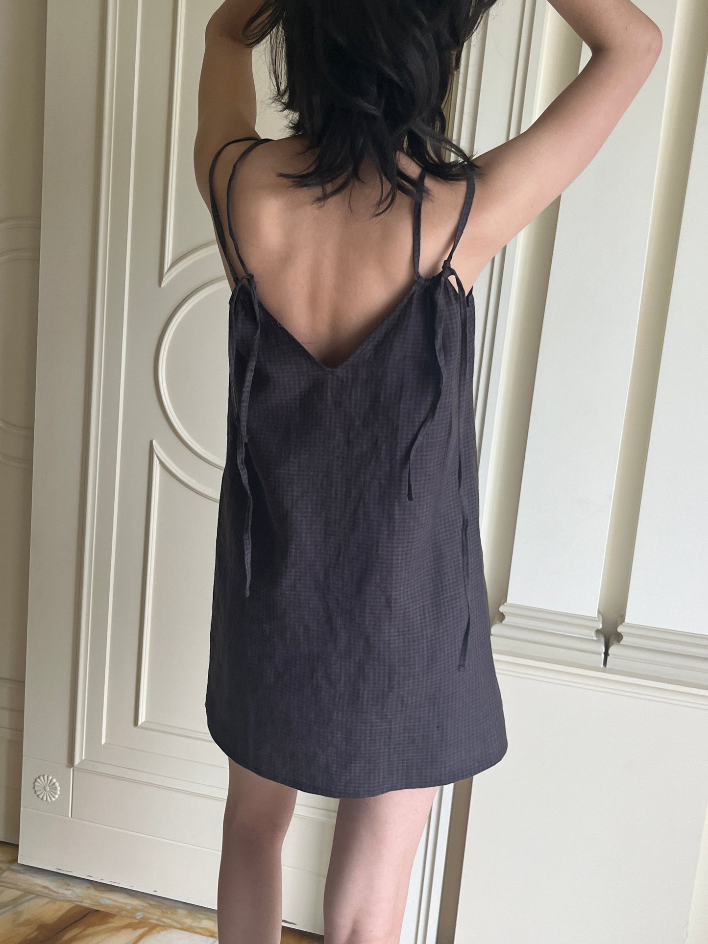 A person with shoulder-length dark hair stands indoors, facing away from the camera, wearing the sleeveless Looped Dress - Slate Check with bow shoulder details and a v neckline. Their arms are raised above their head.