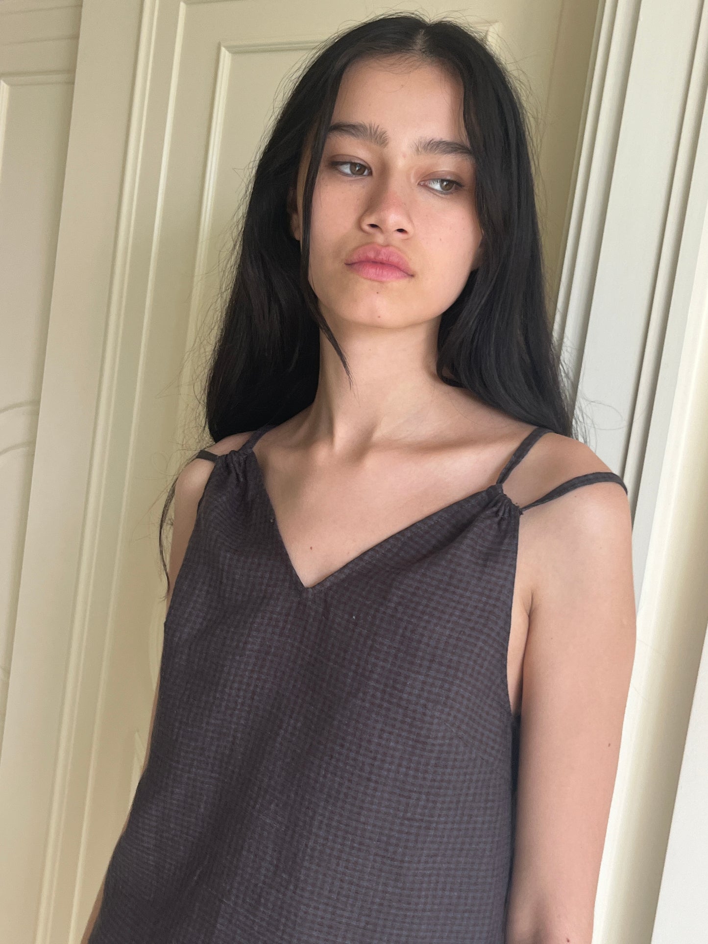A young woman with long dark hair wears the Looped Dress - Slate Check, a sleeveless patterned dress, standing in front of a light-colored door and gazing slightly to the side with a neutral expression.