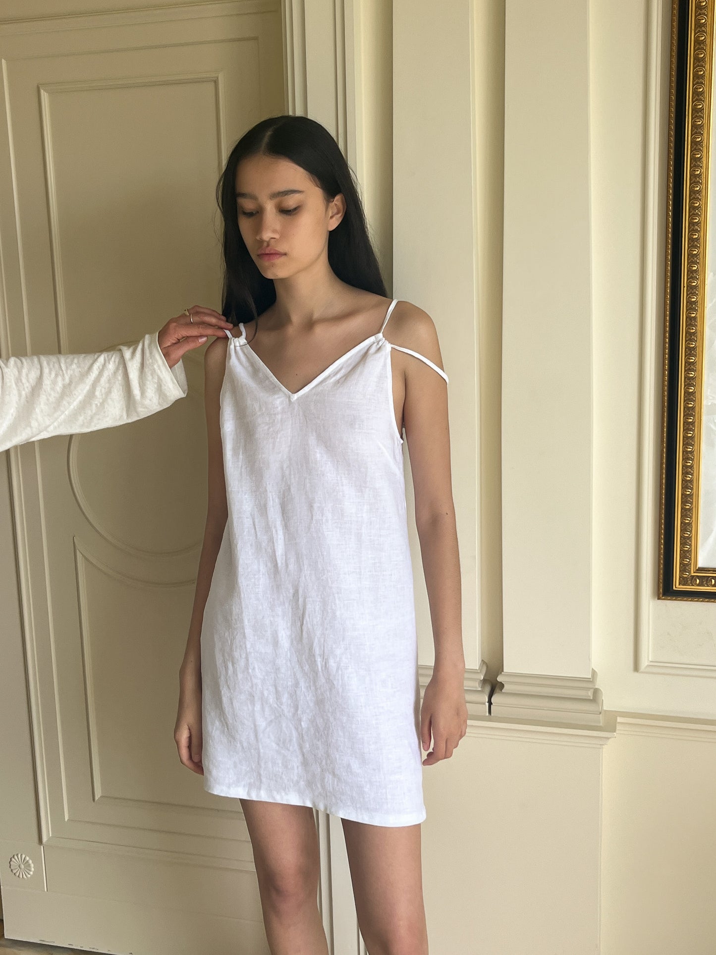 A young woman stands indoors in the Looped Dress - White, a mini-length organic linen piece, while another person adjusts a strap. Cream-colored walls and a decorative gold-framed mirror complete the background.