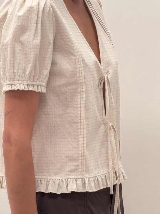 A person wearing the Deiji Studios Pintuck Top - Bone Check, featuring blouson sleeves, a subtle check pattern, ruffled hem, and deep V-neck with tie strings, paired with dark pants.