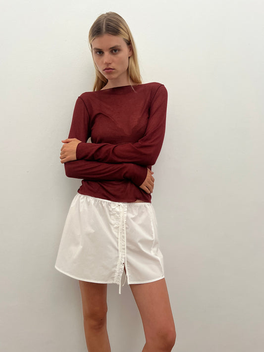 A woman with long blonde hair stands against a white wall, wearing the Knit Long Sleeve - Crimson and a white mini skirt with a front drawstring. Her arms are crossed and she has a neutral expression.