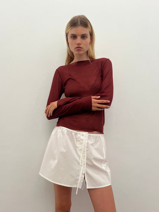 A woman with straight blonde hair stands against a white wall, arms crossed and expression neutral, wearing the Knit Long Sleeve - Crimson and a short white skirt with a front tie detail.