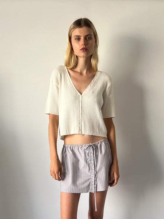 A young woman with straight blonde hair stands against a plain light-colored wall, wearing the One Piece Tee - Chalk and a light gray striped mini skirt with a drawstring.