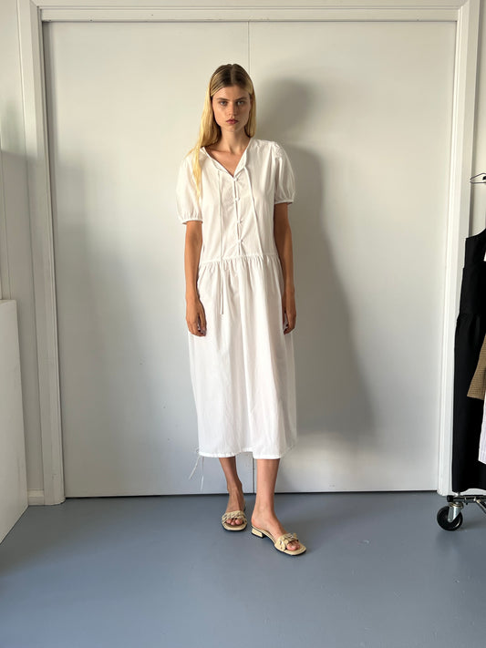 A woman stands indoors against a white wall, wearing the Draw Hem Dress - White with short puff sleeves and beige slide sandals. Her long blonde hair frames her neutral expression. Clothing hangs on a rack to her right.