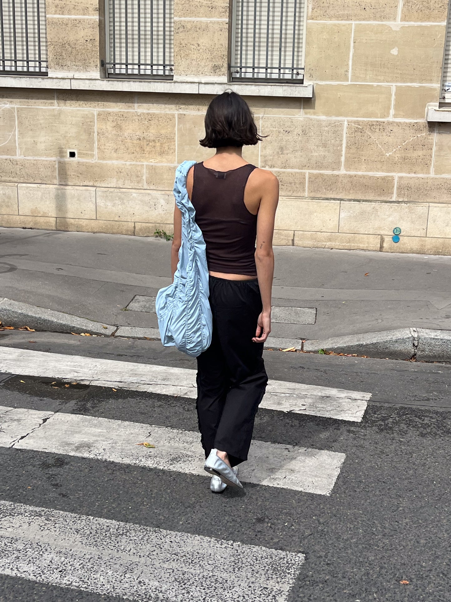 A person with short dark hair, wearing the Knit Tank - Mahogany, black pants, and silver shoes carries a light blue slouchy bag while crossing a street. A beige building with barred windows is visible in the background.