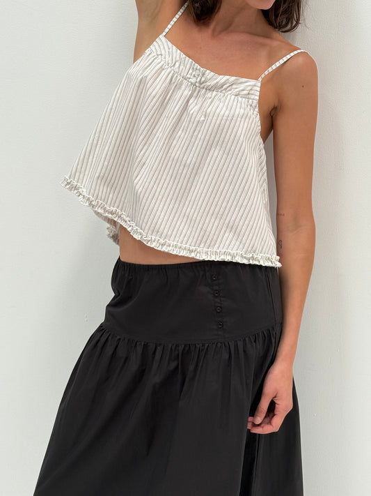 A woman wears a white, loose-fitting, striped spaghetti-strap top with ruffled edges, paired with the Button Skirt - Black by Deiji Studios. She stands against a plain white background.
