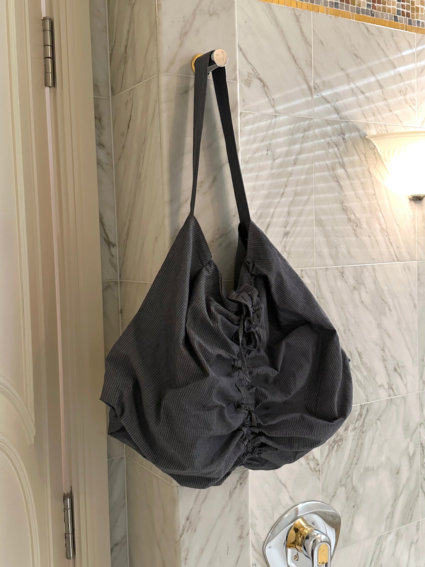 A large blue pinstripe Terrace Bag by Deiji Studios hangs on a round wall hook in a tiled bathroom, near a door and a sconce light.