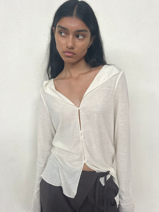Model wearing the Open Long Sleeve Top - Off White by Deiji Studios, made from recycled polyester with a cream ribbed finish, deep V-neckline, and collar, styled with dark brown tie-waist trousers against a white wall.