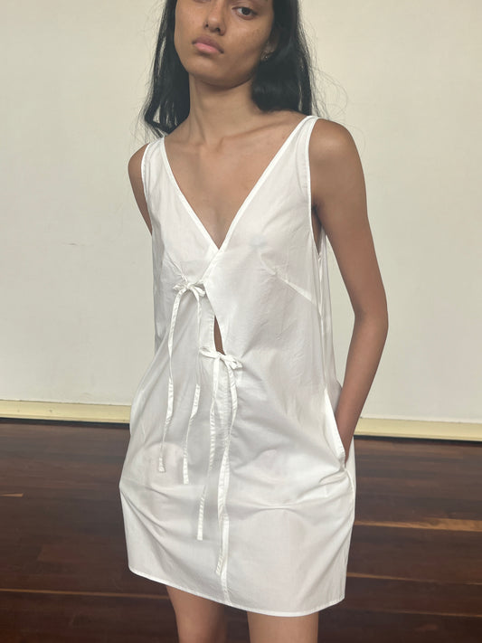 Someone stands indoors on a wooden floor, wearing the Open Tie Mini Dress - White by Deiji Studios, made from organic cotton, with a neutral expression and one hand in their pocket.