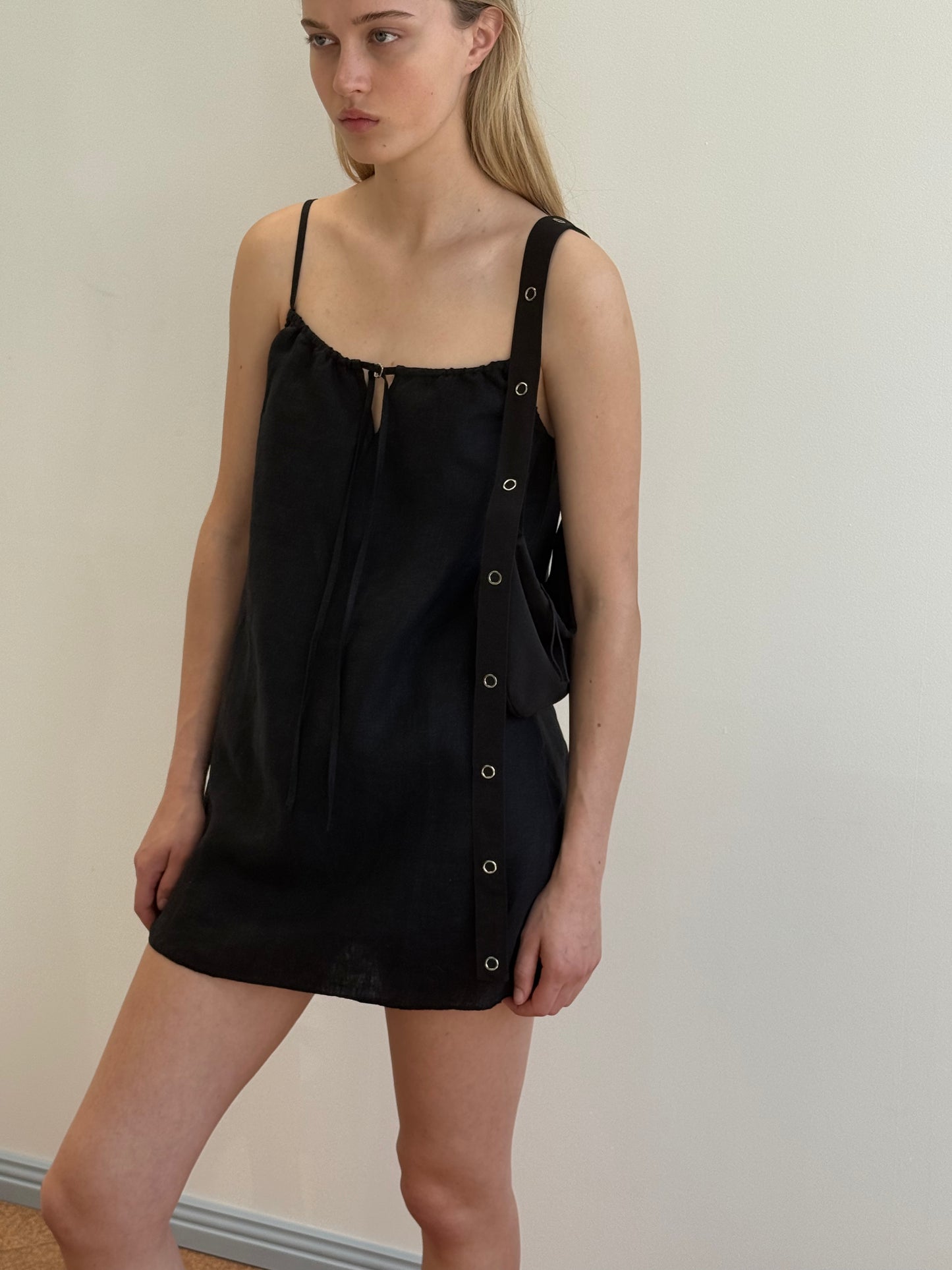 A woman with straight, light hair and a neutral expression stands against a light wall, wearing the Lettuce Mini Dress - Black: a sleeveless French linen dress with thin straps, neckline tie, and large grommets on one side.