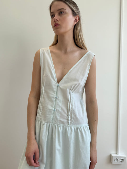 A young woman with long blonde hair wears the sleeveless Deiji Studios Pintuck Dress in Mint, featuring a deep V-neck and button details, standing against a plain light-colored wall.