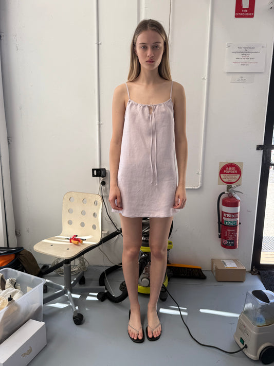 A young woman stands indoors by a white wall wearing the Lettuce Mini Dress - Lilac from Deiji Studios with black flip-flops, near a chair, fire extinguisher, pipes, and various items scattered on the floor.
