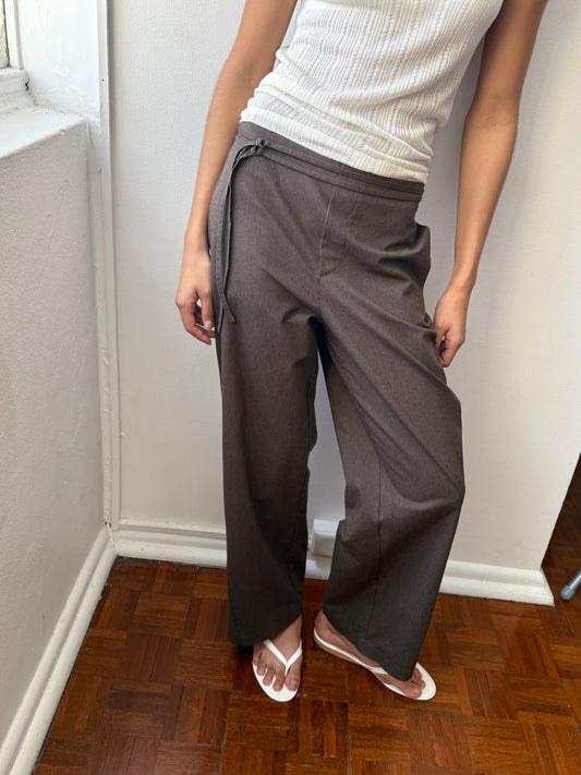 A person stands indoors wearing a white sleeveless ribbed top, Deiji Studios Double Belted Pant - Brown Stripe with a tie waist, and white flip-flops. Only the upper body and lower half of the person are visible.