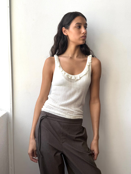 A woman with long, wavy dark hair stands against a white wall, wearing the Neck Ruffle Knit - Off White from Deiji Studios and brown pants, looking to the side with a neutral expression.