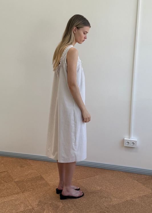 A woman with long hair stands sideways in a simple room, wearing the Button Sweep Dress - Gull Stripe by Deiji Studios and black flat sandals against a beige floor, plain wall, and white electrical outlet.