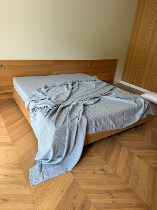 A wooden bed with a headboard sits on a herringbone-patterned floor. The mattress is covered with Sheets - Blue Stripe, which hang over the edge, while a single pillow rests on the floor near a wooden wardrobe.