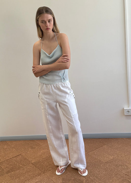 A young woman with straight, light brown hair stands indoors against a plain wall. She wears a light blue camisole top, the Angle Pocket Pant - White, and white sandals. Her arms are crossed and she has a neutral expression.