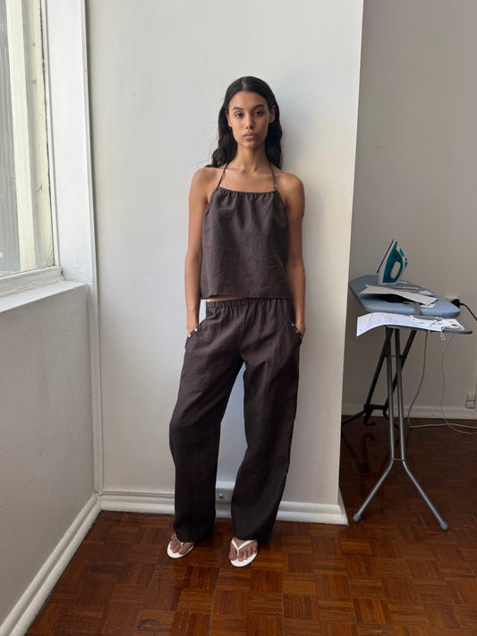 A woman stands indoors by a window, wearing the Deiji Studios O Ring Halter - Bark with matching dark brown wide-leg pants and white sandals. She has her hands in her pockets; an iron and ironing board are in the background.